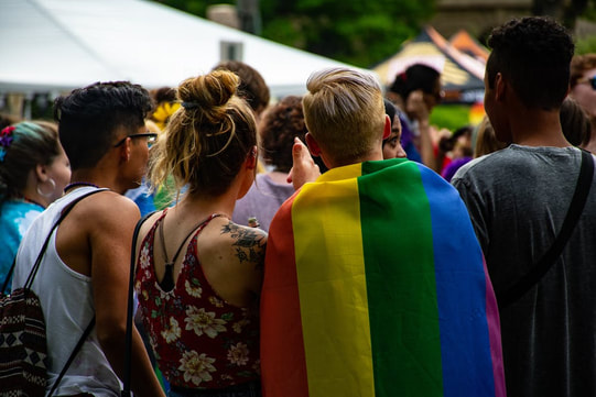 A group of youth are gathered around, one has a rainbow flag draped across their back.