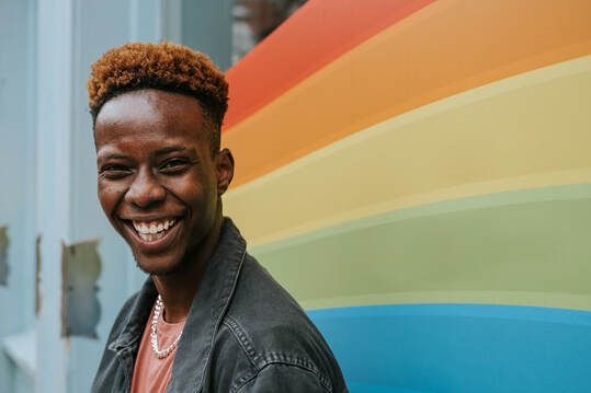 A person with dark skin, orange hair with dark roots, stands in front of a rainbow colored window. They are smiling at the camera.